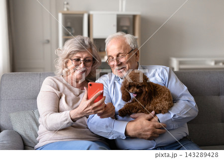 Senior couple checking smartphone relaxing on sofa with poodle dog 143289428