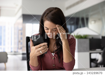 Woman sitting at desk holding smartphone with distressed expression Woman sitting at desk holding smartphone with distressed expression 143289670