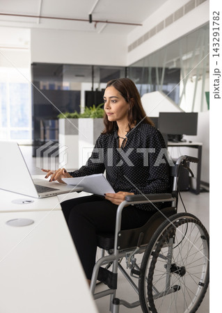 Confident woman in wheelchair using laptop at office desk Confident woman in wheelchair using laptop at office desk 143291782
