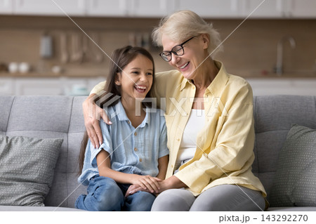 Cheerful grandmother and granddaughter speaking seated on couch Cheerful grandmother and granddaughter speaking seated on couch 143292270
