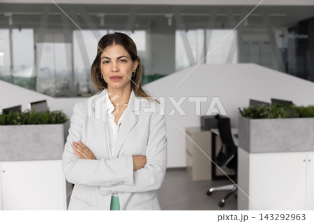 Confident Hispanic female standing with arms crossed in modern office 143292963