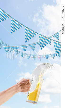 A person holds a large glass beer mug, spilling and splashing beer against a background of bright blue sky with white clouds and traditional Bavarian checkered pennants. Bavarian Oktoberfest Festival 143296176