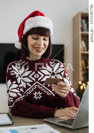 A smiling Caucasian woman wearing a Santa hat and a festive sweater is using a laptop and credit card for online shopping during the holiday season A smiling Caucasian woman wearing a Santa hat and a festive sweater is using a laptop and credit card for online shopping during the holiday season 143296796