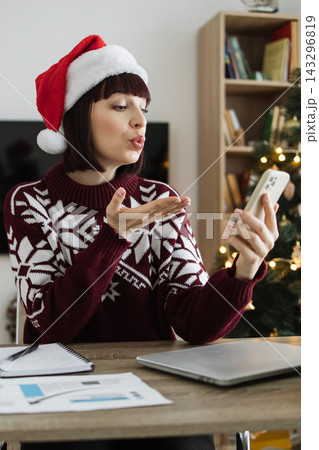 A Caucasian woman wearing a Santa hat and festive sweater blows a kiss while on a video call with her smartphone during the holiday season 143296819