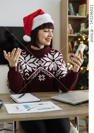 A Caucasian woman wearing a festive Santa hat and a snowflake sweater smiles while video calling on her phone at a desk with a laptop and papers 143296821