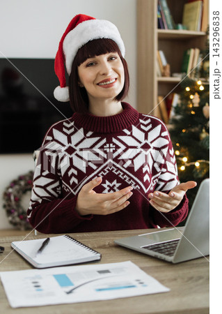 A smiling woman wearing a Santa hat and festive sweater gestures while on a video call with a laptop in front of her 143296838