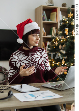 A Caucasian woman wearing a Santa hat and festive sweater is video conferencing on her laptop with a decorated Christmas tree in the background 143296841