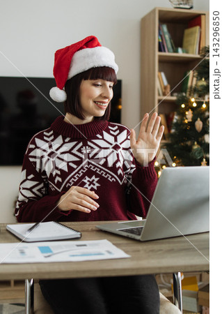 A smiling woman wearing a Santa hat and a festive sweater waves hello during a video call, with a Christmas tree in the background A smiling woman wearing a Santa hat and a festive sweater waves hello during a video call, with a Christmas tree in the background 143296850