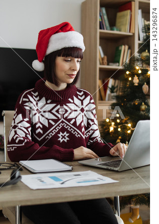 A Caucasian woman wearing a Santa hat and festive sweater works on her laptop at a desk with a Christmas tree in the background A Caucasian woman wearing a Santa hat and festive sweater works on her laptop at a desk with a Christmas tree in the background 143296874