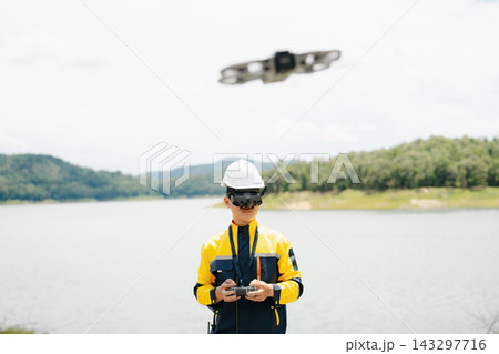 Male engineer in safety gear uses FPV goggles and a remote to pilot a drone. Conducting a high-tech aerial survey near a beautiful natural lake Male engineer in safety gear uses FPV goggles and a remote to pilot a drone. Conducting a high-tech aerial survey near a beautiful natural lake 143297716