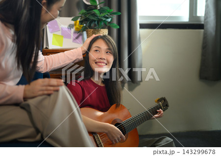 Asian Mother Gently Patting Daughter's Head While She Plays Acoustic Guitar at Home on Mother's Day Asian Mother Gently Patting Daughter's Head While She Plays Acoustic Guitar at Home on Mother's Day 143299678