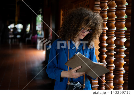 Caucasian Woman Traveler Reading Guidebook at Traditional Wooden Temple Corridor with Camera Caucasian Woman Traveler Reading Guidebook at Traditional Wooden Temple Corridor with Camera 143299713