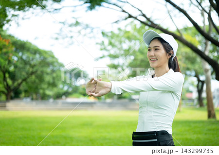 Young Asian Woman Stretching Arms in Green Park Before Workout, Smiling and Happy 143299735