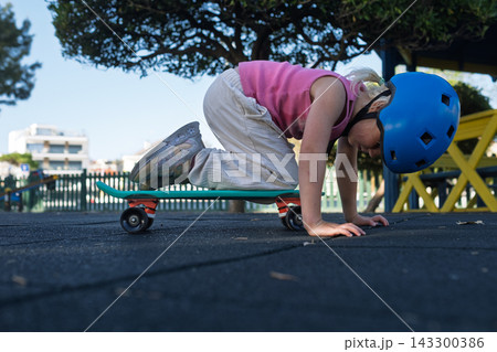 Cute Little Girl in Blue Helmet Sitting on Penny Board at Playground Outdoors 143300386