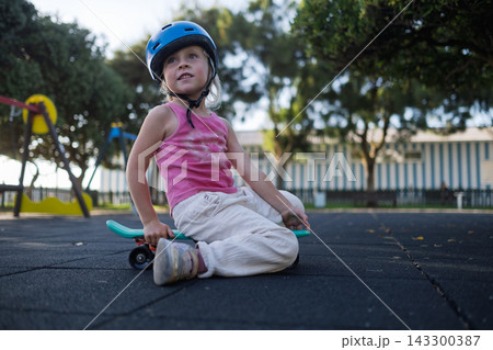 Cute Little Girl in Blue Helmet Sitting on Penny Board at Playground Outdoors 143300387