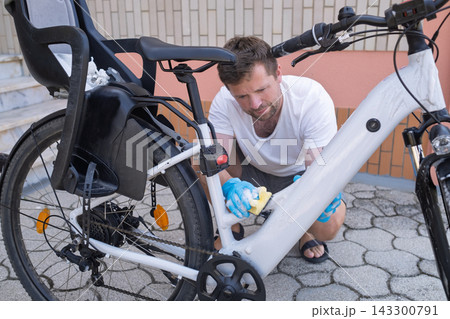 man wearing blue rubber gloves washing a white electric bicycle using a soapy sponge outdoors. man wearing blue rubber gloves washing a white electric bicycle using a soapy sponge outdoors. 143300791