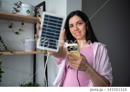 Young woman holding portable solar panel and smartphone in a home interior. Solar charging sustainable living and renewable energy technology 143301328
