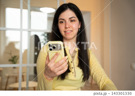 Young woman holding smartphone during a casual video call in a home interior. Digital communication social connection and mobile networking Young woman holding smartphone during a casual video call in a home interior. Digital communication social connection and mobile networking 143301330