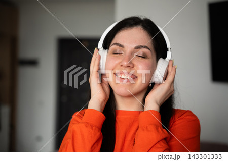 Smiling woman wearing white over-ear headphones with eyes closed at home. Personal music listening relaxation self-care and entertainment Smiling woman wearing white over-ear headphones with eyes closed at home. Personal music listening relaxation self-care and entertainment 143301333