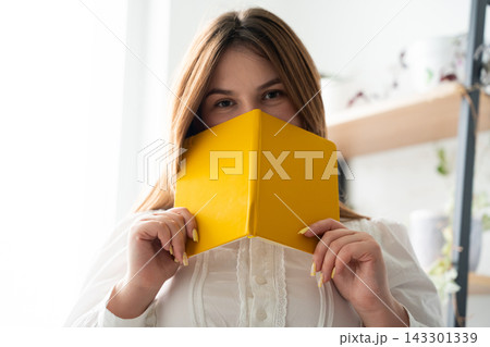 Young woman holding a yellow notebook over her face in a bright home interior. Personal diary privacy creative learning and stationery 143301339