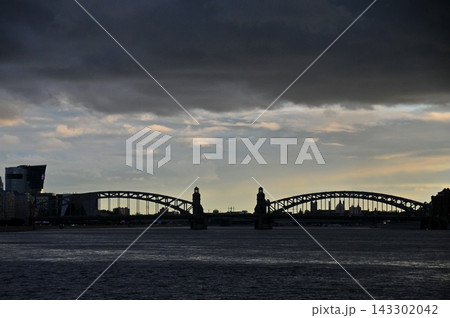 Silhouetted steel arch bridge spanning wide river under dramatic cloudy sky at dusk with city skyline in background urban infrastructure Silhouetted steel arch bridge spanning wide river under dramatic cloudy sky at dusk with city skyline in background urban infrastructure 143302042