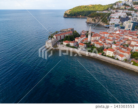 Budva Montenegro historic center occupies walled peninsula along Adriatic coast. Stone buildings with terracotta roofs cluster around church tower, with modern resorts along nearby shore. Budva Montenegro historic center occupies walled peninsula along Adriatic coast. Stone buildings with terracotta roofs cluster around church tower, with modern resorts along nearby shore. 143303553