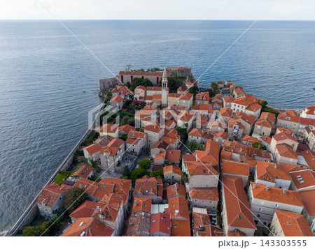 Budva Montenegro sits beside Adriatic Sea, its old town clustered beneath tall church tower. Stone houses with terracotta roofs fill fortified peninsula, seen from elevated coastal viewpoint. Budva Montenegro sits beside Adriatic Sea, its old town clustered beneath tall church tower. Stone houses with terracotta roofs fill fortified peninsula, seen from elevated coastal viewpoint. 143303555