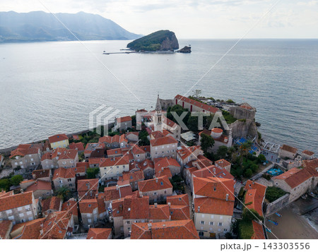 Budva Montenegro old town rests beside calm Adriatic waters, its dense stone houses topped with red tiles. Aerial panorama includes fortified waterfront, church spire and green island offshore. Budva Montenegro old town rests beside calm Adriatic waters, its dense stone houses topped with red tiles. Aerial panorama includes fortified waterfront, church spire and green island offshore. 143303556