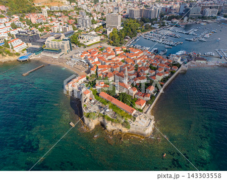 Budva Montenegros coastal old town fills fortified peninsula, ringed by clear Adriatic water. High aerial perspective reveals terracotta roofs, harbor boats, sandy beaches and modern hillside blocks. 143303558