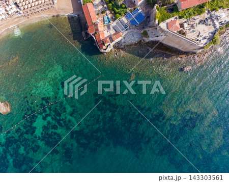 Budva Montenegro shoreline meets transparent Adriatic water, where swimmers are visible beside floating safety line. Overhead coastal view reveals stone walls, red tiled roofs and beach umbrellas. 143303561