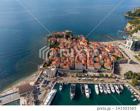 Budva Montenegro old town sits within stone walls beside Adriatic Sea. Terracotta rooftops cluster above harbor yachts, with beaches, cliffs and coastal buildings completing sunlit view. Budva Montenegro old town sits within stone walls beside Adriatic Sea. Terracotta rooftops cluster above harbor yachts, with beaches, cliffs and coastal buildings completing sunlit view. 143303565