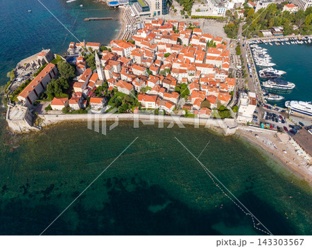Budva Montenegro old town fills coastal peninsula inside ancient stone ramparts. Red roofed buildings overlook Adriatic water, while moored yachts and beach shoreline mark this scenic seaside setting. 143303567