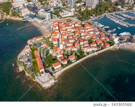 Budva Montenegro old town rests on coastal peninsula, its terracotta roofs enclosed by stone walls beside Adriatic water. Aerial view includes marina packed with boats and buildings. 143303568