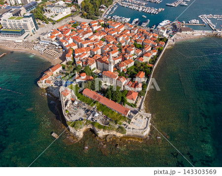 Budva Montenegro appears from above, with historic Old Town packed across coastal peninsula. Stone ramparts, terracotta roofs and turquoise Adriatic water frame harbor and boats along its shoreline. Budva Montenegro appears from above, with historic Old Town packed across coastal peninsula. Stone ramparts, terracotta roofs and turquoise Adriatic water frame harbor and boats along its shoreline. 143303569