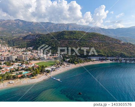 Budva Montenegro sits between mountain slopes and Adriatic Sea, with long sandy beach edging waterfront. Coastal neighborhoods and bright turquoise shallows shape this resort panorama. 143303576