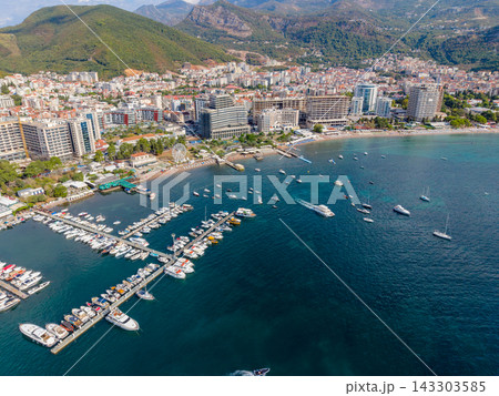 Budva Montenegro appears from above, with crowded marina and boats set against citys beachfront skyline. Deep blue Adriatic water meets urban buildings beneath wooded mountains, while boats fill 143303585
