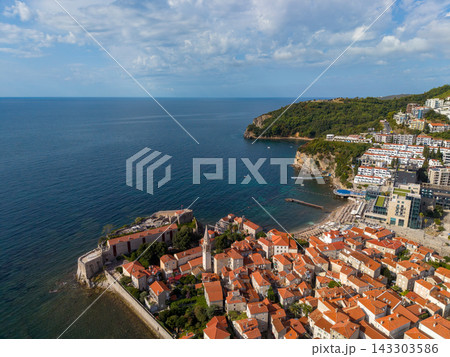 Budva Montenegro appears above Adriatic, where old town rooftops cluster within coastal fortifications. Sheltered bay, green headland and contemporary seaside hotels complete sunny panorama. 143303586