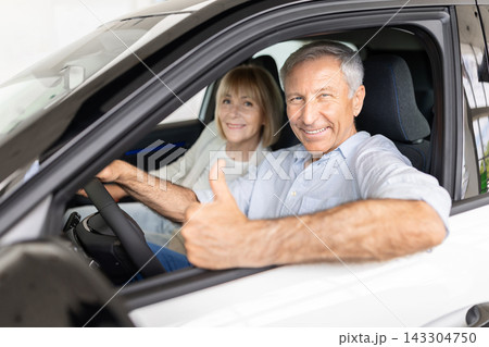 Smiling senior couple sitting inside car at dealership showroom, showing thumb up 143304750