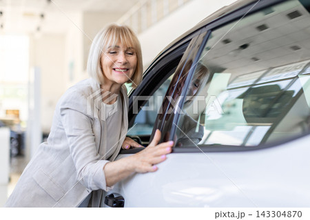 Senior woman closely examining exterior and window of vehicle in dealership showroom Senior woman closely examining exterior and window of vehicle in dealership showroom 143304870