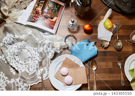 Elegant vintage dining table scene featuring lace tablecloth, open art book displaying classical painting, fresh fruit, glassware, candlelight, and ornate silverware on wooden surface 143305550