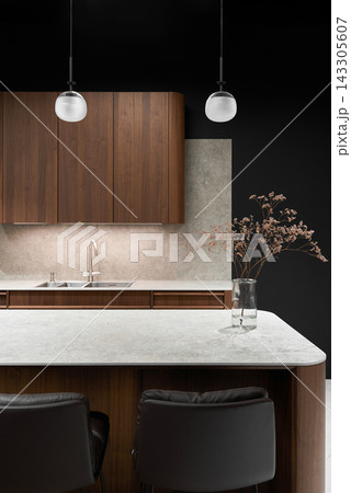 Modern kitchen interior with wood cabinets, dark concrete backsplash, marble countertop, glass pendant lights, double sink, and vase with dried blossoms under warm lighting 143305607