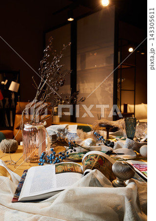 Cozy indoor still life with open magazine, pink glass vase holding dried branches, blue berries, ceramics, and textiles on wooden table under warm ambient lighting Cozy indoor still life with open magazine, pink glass vase holding dried branches, blue berries, ceramics, and textiles on wooden table under warm ambient lighting 143305611