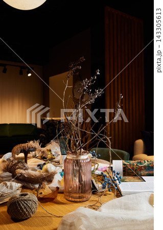 Dried twig arrangement in ribbed glass vase on cluttered wooden table with craft supplies, warm indoor lighting and moody vertical wood panel background 143305613