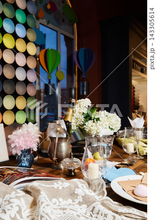 Colorful party decor scene with round wall discs, hanging paper balloons, champagne bottle in ice, white hydrangea centerpiece, candles, and elegant table setting on wooden background Colorful party decor scene with round wall discs, hanging paper balloons, champagne bottle in ice, white hydrangea centerpiece, candles, and elegant table setting on wooden background 143305614