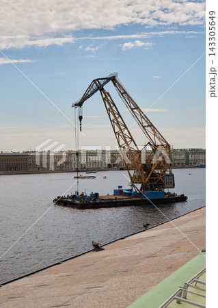 Rusty industrial lattice boom crane on barge lifting cargo over river with hook and cables and classical city skyline under blue sky Rusty industrial lattice boom crane on barge lifting cargo over river with hook and cables and classical city skyline under blue sky 143305649