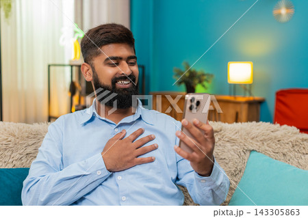 Indian man smiles gently hand on heart viewing touching nostalgic photo on smartphone memory indoors 143305863