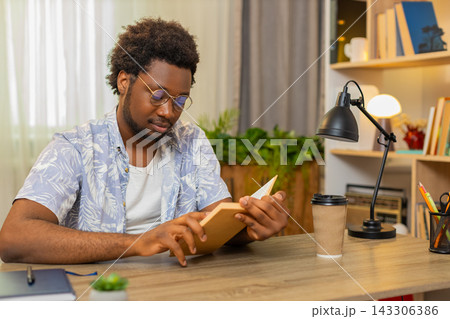 African American young man on home table reads a book and enjoys calm leisure time during relaxation African American young man on home table reads a book and enjoys calm leisure time during relaxation 143306386