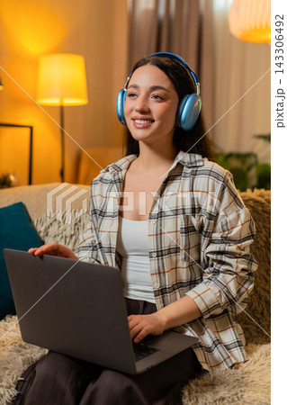 Young woman on home sofa puts on headphones and starts working on laptop in cozy living room at home 143306492