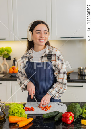 Young woman cutting cherry tomatoes listening to music preparing fresh vegetarian salad at home meal Young woman cutting cherry tomatoes listening to music preparing fresh vegetarian salad at home meal 143306507