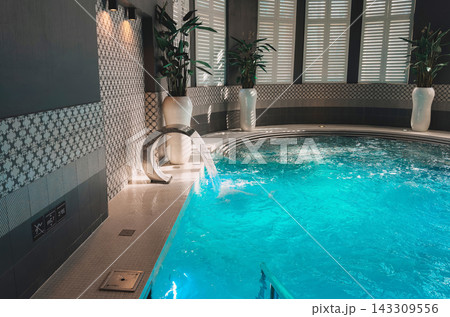 uxurious and modern indoor hydrotherapy pool featuring sparkling turquoise water, a cascading water jet feature, decorative patterned tile walls, and elegant white planters with green plants, creating uxurious and modern indoor hydrotherapy pool featuring sparkling turquoise water, a cascading water jet feature, decorative patterned tile walls, and elegant white planters with green plants, creating 143309556